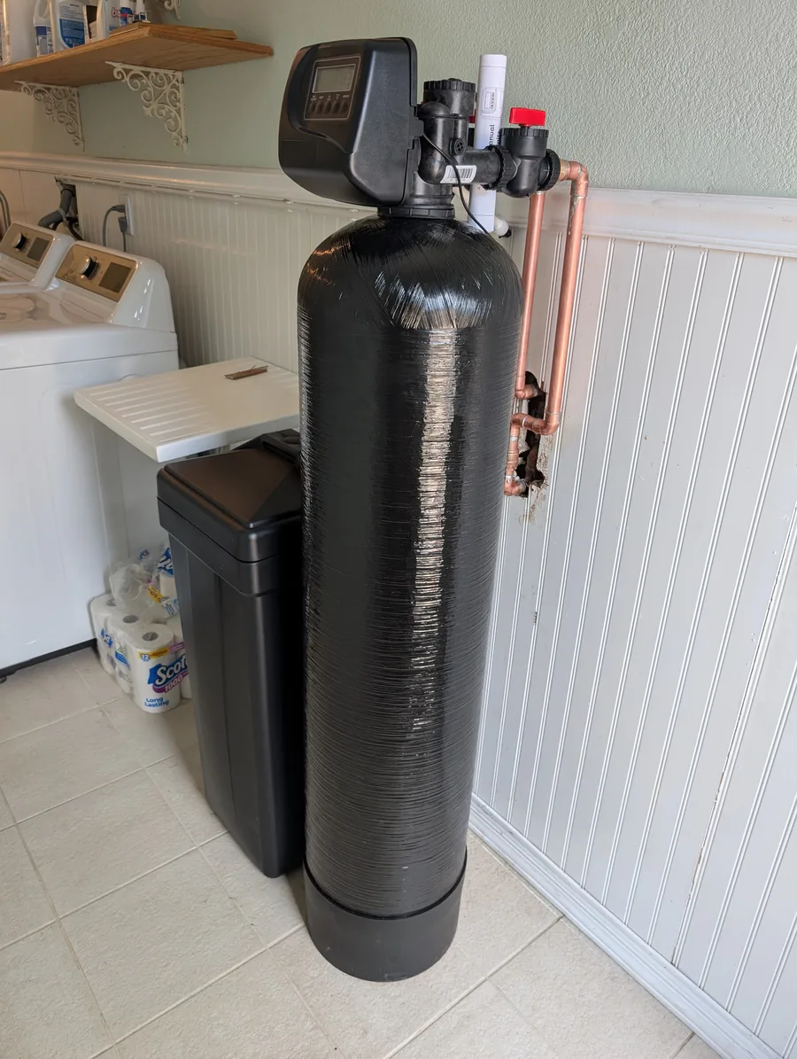 Water softener tank with its control valve and copper connections, brine tank beside it, in a laundry room